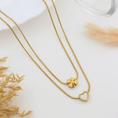 Two delicate gold-tone necklaces on a white background, one featuring a small four-leaf clover pendant and the other a hollow heart pendant, styled with dried grass and a glass rim.