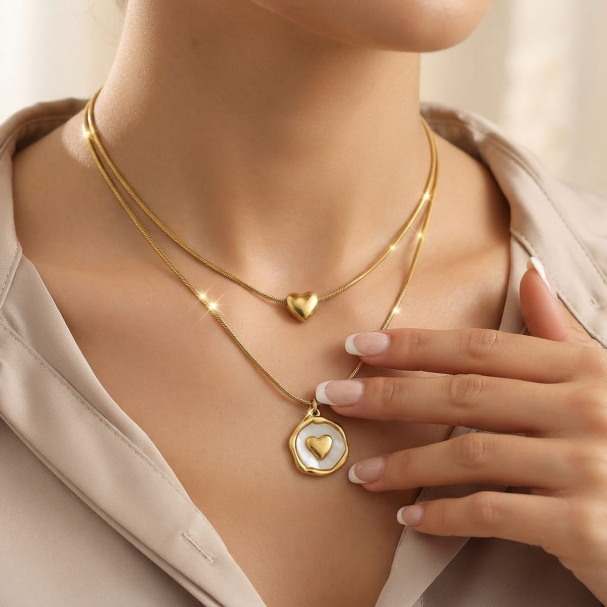 A close-up of a model wearing a double-layered gold necklace with a small heart pendant on the top layer and a larger oval pendant with a heart in the center on the bottom layer. The model is wearing a beige satin shirt.