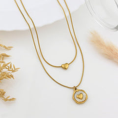 A gold-toned double-strand necklace with a small heart pendant on one chain and a larger open circle pendant with a smaller heart in the center on the other chain, styled on a white surface with dried botanicals and a glass rim.