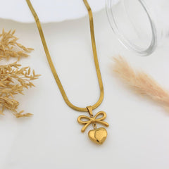 A gold snake chain necklace with a bow and heart pendant, styled on a white surface with dried grass and a glass prop.