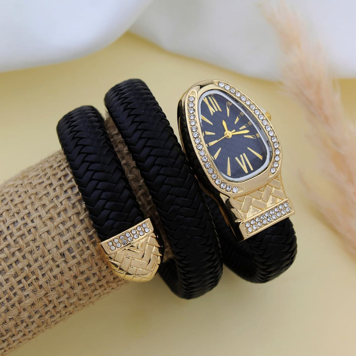 A black braided wrap-style watch with a gold-tone case, crystal-studded bezel, and dark blue dial, displayed on a burlap-wrapped wrist against a soft cream background.