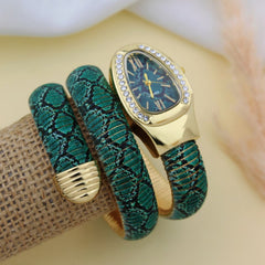 A gold-tone watch with a blue dial and crystal-studded bezel, displayed on a textured green and black snake-print wrap bracelet with a gold clasp, resting on a burlap-wrapped stand against a soft beige background.