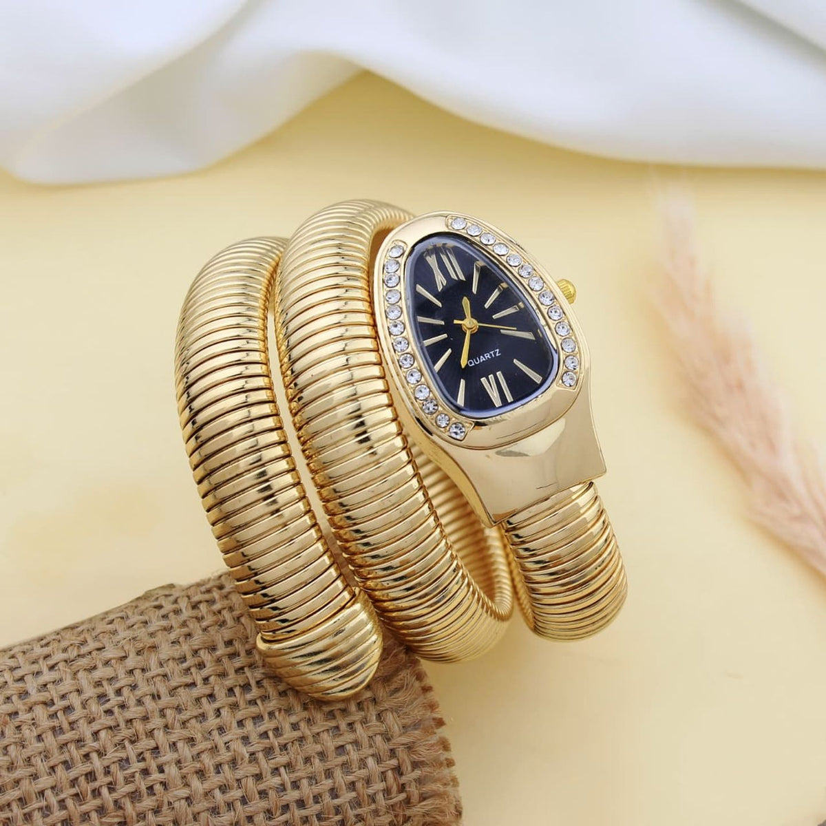 A gold-plated bangle watch with a coiled snake-like strap, a black dial, and crystal accents around the bezel, displayed on a beige surface with a white fabric and burlap prop.