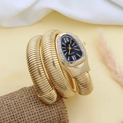 A gold-plated bangle watch with a coiled snake-like strap, a black dial, and crystal accents around the bezel, displayed on a beige surface with a white fabric and burlap prop.