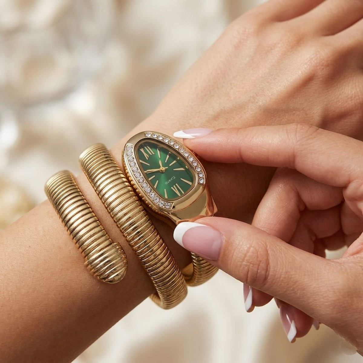 A close-up lifestyle shot of a woman's wrist wearing a gold-tone watch with a green dial and crystal-embellished bezel, paired with a matching gold bangle bracelet.