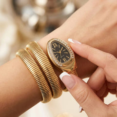 A close-up lifestyle shot of a woman's wrist wearing a gold-plated watch with a black dial and crystal-embellished bezel, paired with a matching gold snake-bangle bracelet.