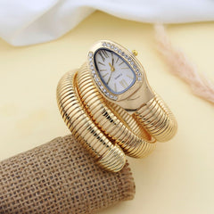 A gold-tone snake-style wristwatch with a rhinestone-accented oval bezel and white dial, displayed on a burlap-textured prop against a soft cream background.