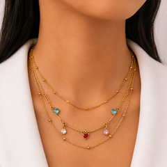 A close-up of a model wearing a gold multi-layered necklace with small bead details and heart-shaped charms in red, blue, and pink, styled with a white blazer.