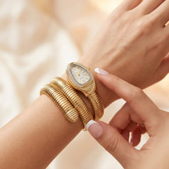 A gold snake-wrap bracelet watch with a bezel-set crystal crown, worn on a wrist alongside a matching gold bangle bracelet, shown on a model's wrist against a soft beige background.
