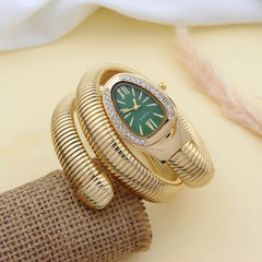A gold-plated bangle watch with a green dial and crystal-embellished bezel, displayed on a textured burlap roll against a soft beige background.