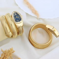 Gold-tone snake-style jewelry set including a wrap bracelet watch with a blue dial and a matching snake ring, styled on white fabric with dried grass accents.