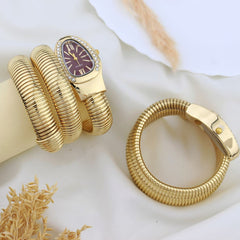 A gold-toned snake-style watch with a purple dial and crystal bezel, displayed alongside a matching gold bangle bracelet on a soft white fabric background with dried grass accents.