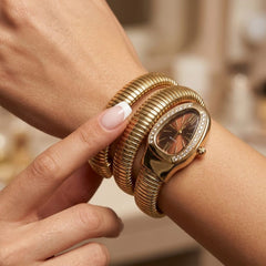 A close-up lifestyle shot of a woman's wrist wearing a gold-plated multi-band watch with a snake-skin textured strap and a diamond-accented oval bezel, alongside stacked gold bangles.