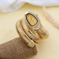Gold-tone snake-style bangle watch with a rectangular crystal-accented dial, displayed on a burlap-like prop against a soft cream background.