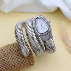 A silver-toned bangle watch with a coiled snake-like strap and an oval white dial accented by small rhinestones, displayed on a beige surface with a burlap roll and soft fabric in the background.