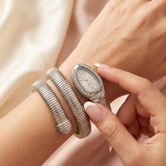 A close-up lifestyle shot of a silver-toned snake-style bangle watch with an oval face and crystal embellishments, worn on a wrist and gently touched by another hand.