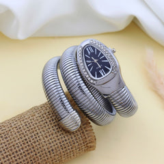 A silver-tone snake-style bangle watch with a dark navy dial and crystal rhinestone bezel, displayed on a burlap roll with a soft cream fabric background.