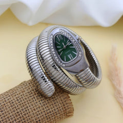 A silver-toned snake-style cuff watch with a green dial and crystal embellishments around the bezel, displayed on a burlap block against a soft beige background with white fabric.