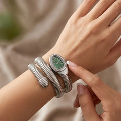 A close-up of a woman's wrist wearing a silver multi-band watch with a green dial and crystal-encrusted bezel, alongside a thick silver snake-style bangle, against a soft beige fabric background.