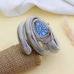 A silver-toned coiled bangle watch with a blue dial and crystal accents, displayed on a burlap-wrapped prop against a soft cream background.