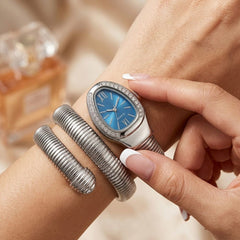 A close-up lifestyle shot of a woman's wrist wearing a silver-tone watch with a blue dial and crystal-embellished bezel, alongside a matching silver bangle bracelet.