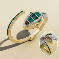 Gold cuff bracelet featuring a snake head motif with emerald-cut green gemstones and clear crystal accents, shown on a beige background with a circular inset of the watch face side.