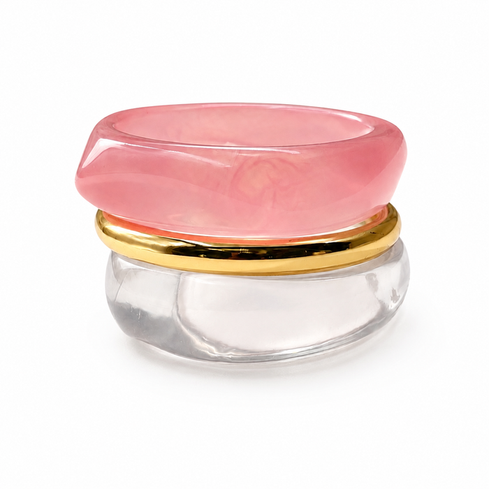 A modern stacked ring featuring a thick, smooth pink stone top, a polished gold band, and a clear glass or acrylic base, shown on a white background.