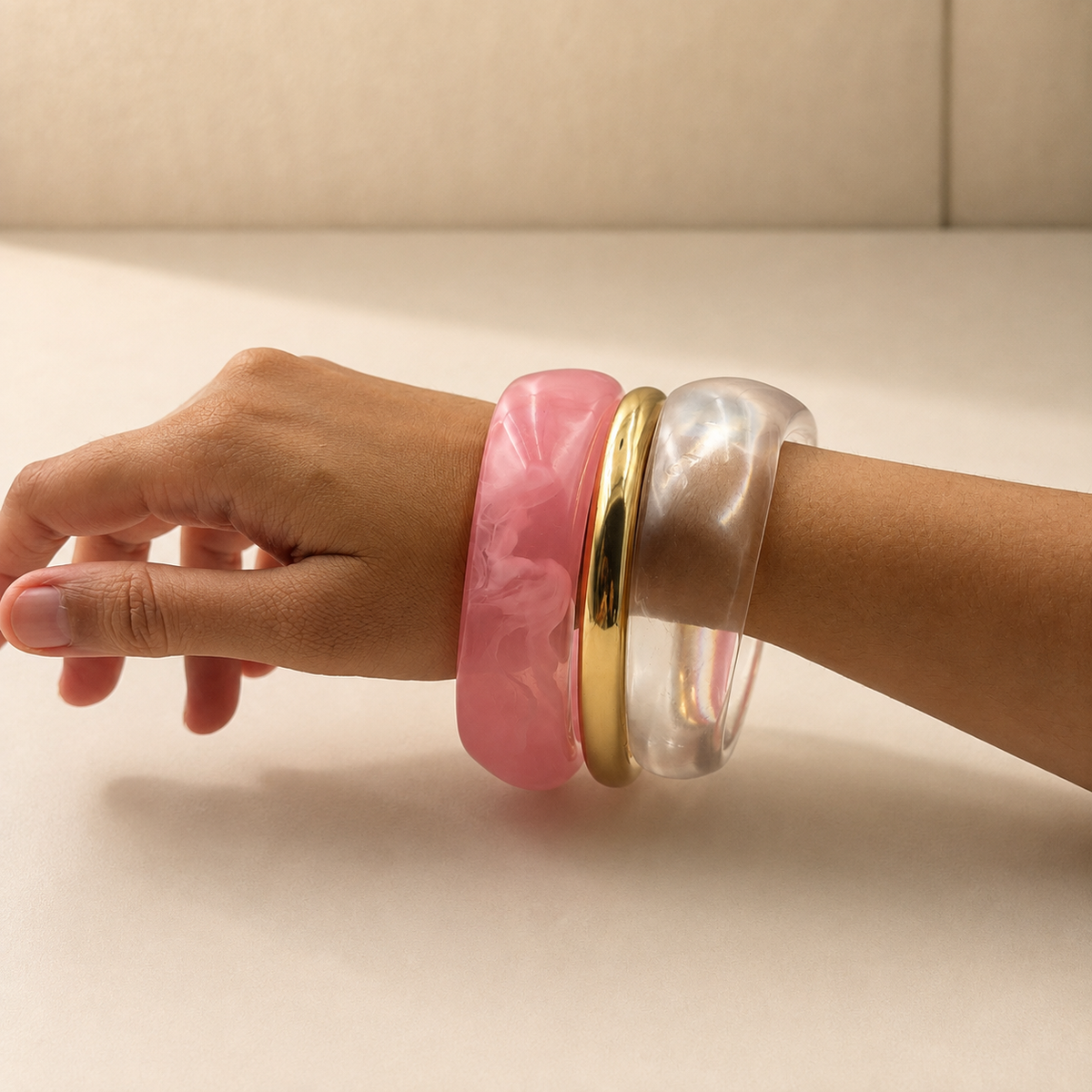 Three wide bangles worn on a person's wrist: a thick pink marbled bangle, a slim polished gold bangle, and a translucent clear bangle, shown against a neutral beige background.