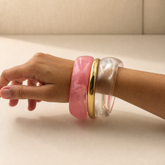 Three wide bangles worn on a person's wrist: a thick pink marbled bangle, a slim polished gold bangle, and a translucent clear bangle, shown against a neutral beige background.