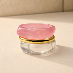 A pink translucent stone ring with a polished gold band and a clear glass base, photographed on a warm beige surface with soft shadows.