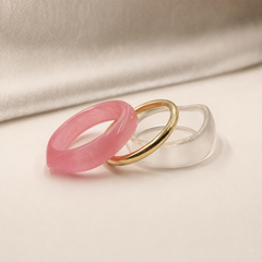 Three minimalist band rings arranged in a stack on a light beige surface with a draped cream fabric backdrop. The front ring is translucent pink, the middle ring is polished gold, and the back ring is clear.
