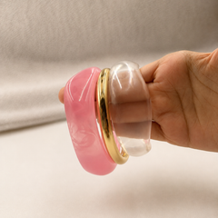 A hand holding a set of three stacked bangles: a thick pink bangle, a slim gold bangle, and a clear bangle, against a neutral light gray fabric background.