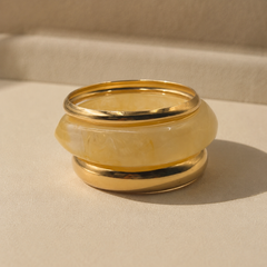 A gold-plated metal ring with a central, rounded translucent pale yellow inlay, photographed on a neutral beige textured surface with a soft shadow.