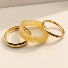 Three minimalist rings on a light beige surface: two polished yellow gold rings and one smooth translucent pale yellow stone ring, arranged in an overlapping composition.