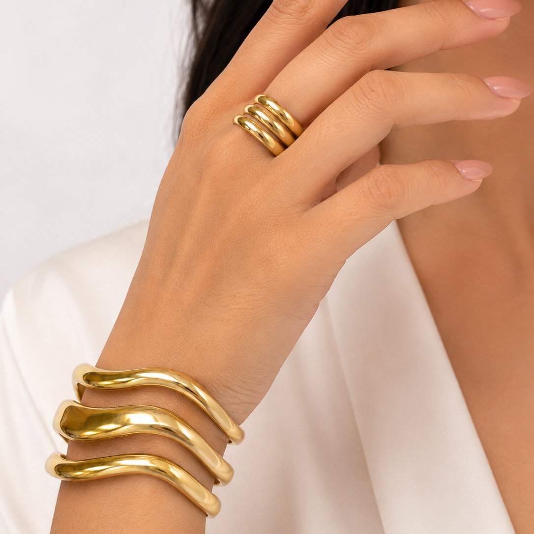 A close-up of a person's hand wearing three slim gold rings on the fingers and a wide, wavy gold cuff bracelet on the wrist, against a soft light background.