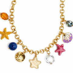 Gold-tone chain necklace with sea-themed charms including two starfish, a pearl shell charm, a blue bead, a faceted amber bead, a gold medallion, and a clear faceted crystal charm, shown on a white background.