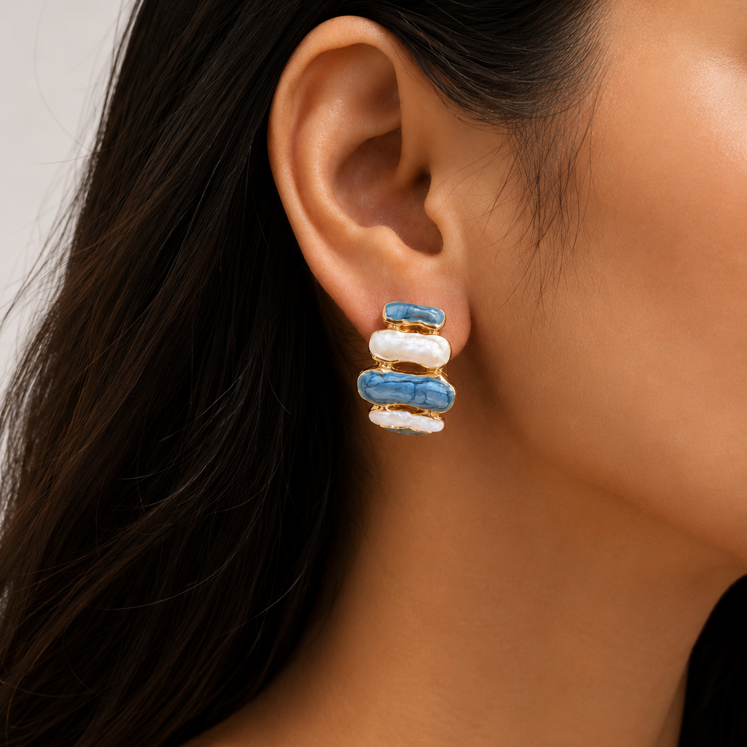 A close-up of a model's ear wearing a small stacked earring with blue and white enamel-like segments outlined in gold.