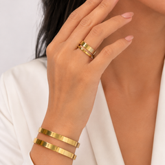 A close-up of a woman's hand and wrist wearing two gold rings and two matching gold bangles, styled with a white blazer against a light gray background.