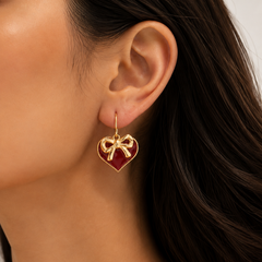 A close-up of a model's ear wearing a gold heart-shaped dangle earring with a red enamel center and a gold bow motif.