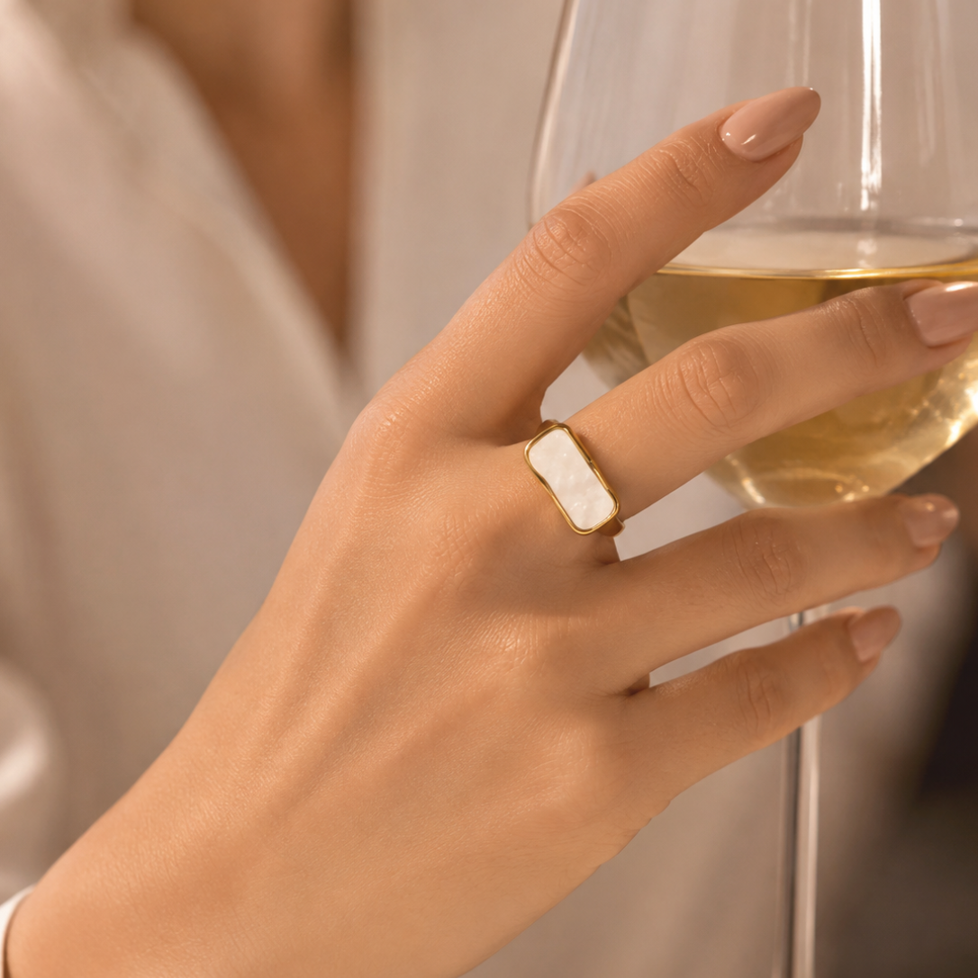A close-up of a hand holding a glass of white wine, wearing a gold ring with a white mother-of-pearl rectangular inlay.