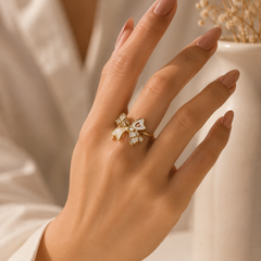 A close-up of a hand wearing a delicate gold ring with a bow-shaped setting and small clear stones, styled with a white robe and a white vase in the background.