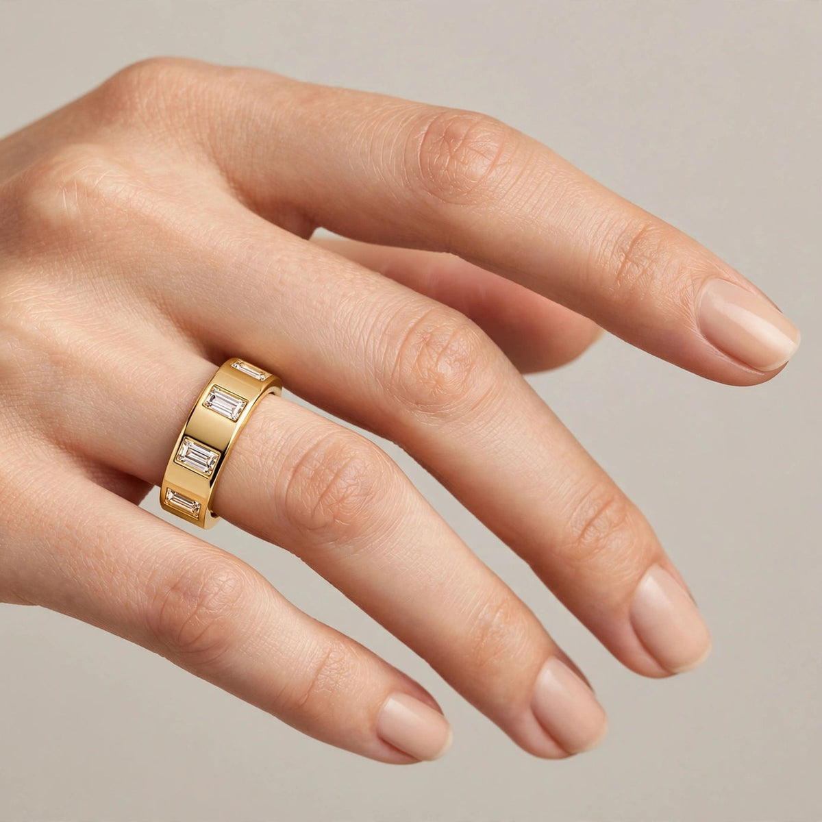 A close-up of a hand wearing a yellow gold band ring with rectangular clear baguette and round diamonds, set against a soft neutral beige background.