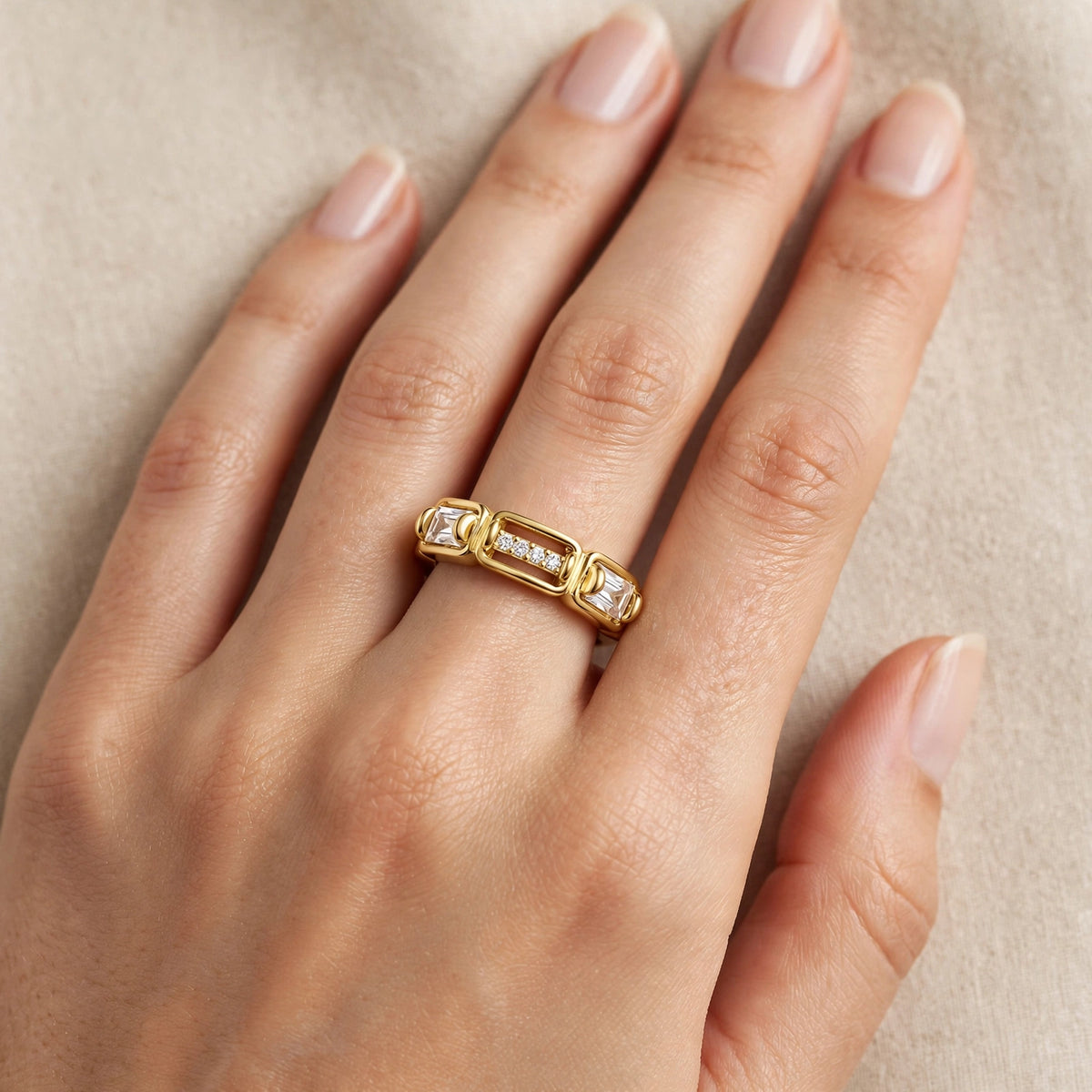 A close-up of a hand wearing a gold ring with small clear gemstones set in a rectangular band, photographed against a soft beige background.