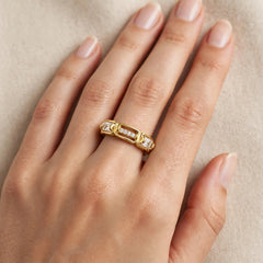 A close-up of a hand wearing a gold ring with small clear gemstones set in a rectangular band, photographed against a soft beige background.