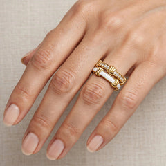 A close-up of a hand wearing two gold rings, including a distinctive ring with a white rectangular inlay and a textured gold band, set against a neutral beige textured background.