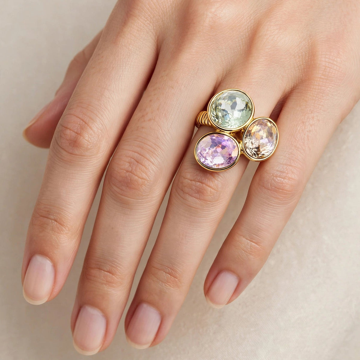 A close-up of a hand wearing a gold ring with three oval gemstones in pale green, lavender purple, and pink.