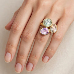 A close-up of a hand wearing a gold ring with three oval gemstones in pale green, lavender purple, and pink.