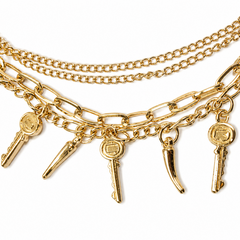 A gold-toned layered chain necklace featuring elongated paperclip links, spiked bar charms, and key-shaped pendants with circular medallions, displayed on a clean white background.