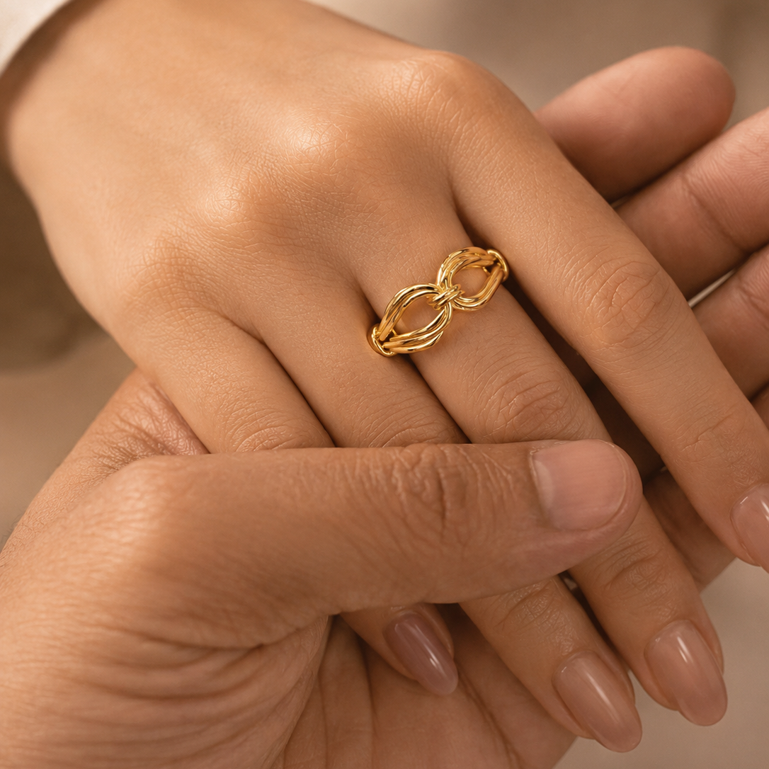 A close-up lifestyle shot of a gold infinity-style ring worn on a finger, held gently by another hand against a soft beige background.