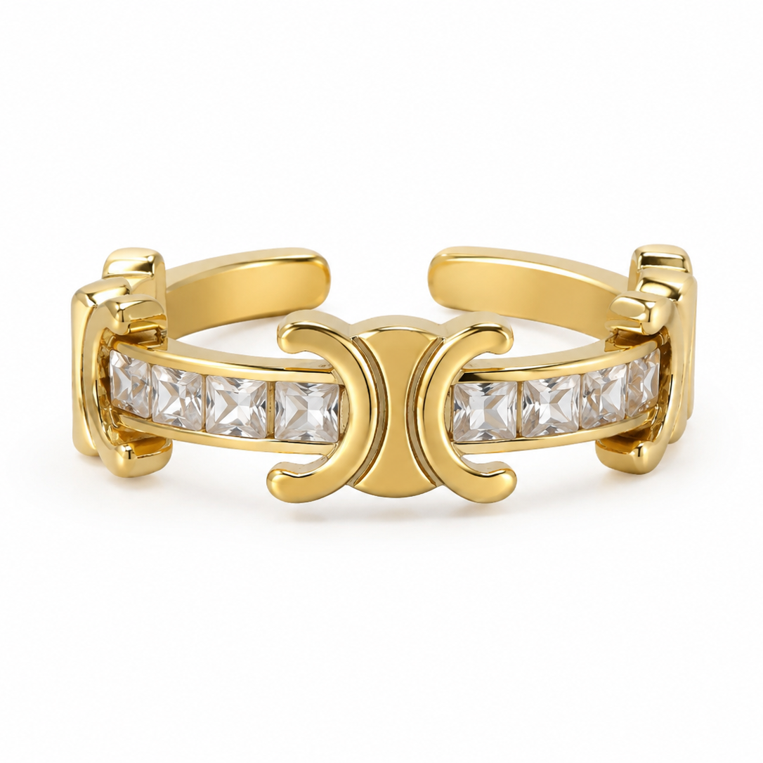 A gold-tone ring featuring a central channel set with clear square-cut stones and a prominent crescent-shaped centerpiece, photographed on a clean white background.